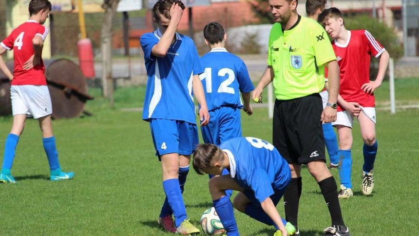 Jogadores de futebol em campo com árbitro. Um deles está no chão, próximo à bola.