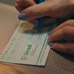 Mãos preenchendo cheque do Sicredi com caneta azul. Foco na escrita 'Dois mil'.