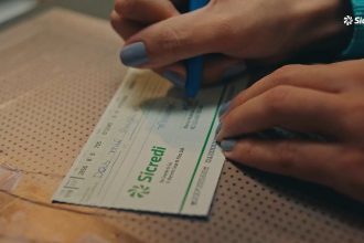 Mãos preenchendo cheque do Sicredi com caneta azul. Foco na escrita 'Dois mil'.