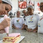 Mulher decora bolo com glacê rosa, com outras mulheres observando em um curso do Senac.