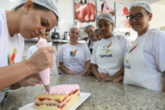 Mulher decora bolo com glacê rosa, com outras mulheres observando em um curso do Senac.