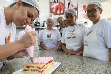 Mulher decora bolo com glacê rosa, com outras mulheres observando em um curso do Senac.