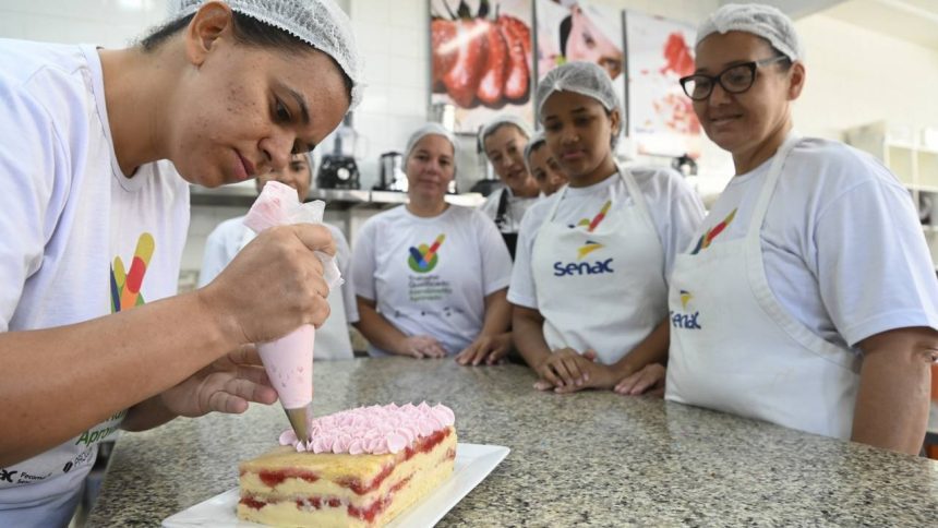 Mulher decora bolo com glacê rosa, com outras mulheres observando em um curso do Senac.
