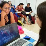 Mulher sorrindo com óculos, olhando para outra pessoa, com um laptop exibindo gráficos ambientais em primeiro plano.