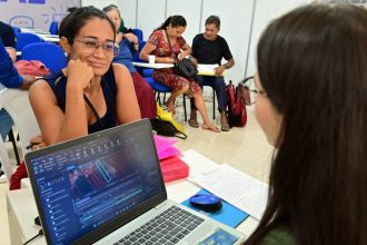 Mulher sorrindo com óculos, olhando para outra pessoa, com um laptop exibindo gráficos ambientais em primeiro plano.