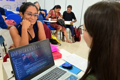 Mulher sorrindo com óculos, olhando para outra pessoa, com um laptop exibindo gráficos ambientais em primeiro plano.