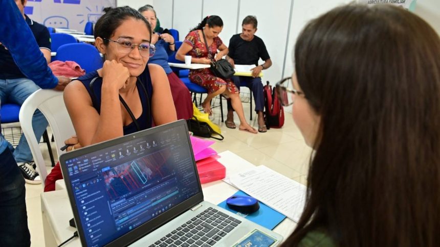 Mulher sorrindo com óculos, olhando para outra pessoa, com um laptop exibindo gráficos ambientais em primeiro plano.