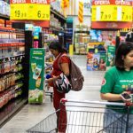 Mulheres comprando em supermercado, com promoções de produtos. Uma delas está grávida e a outra com carrinho de compras.
