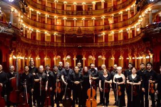 Orquestra de Câmara em palco, com músicos segurando instrumentos de corda, em teatro clássico.