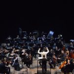 Orquestra sinfônica no palco, com maestro conduzindo e músicos tocando instrumentos.
