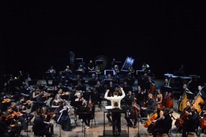 Orquestra sinfônica no palco, com maestro conduzindo e músicos tocando instrumentos.