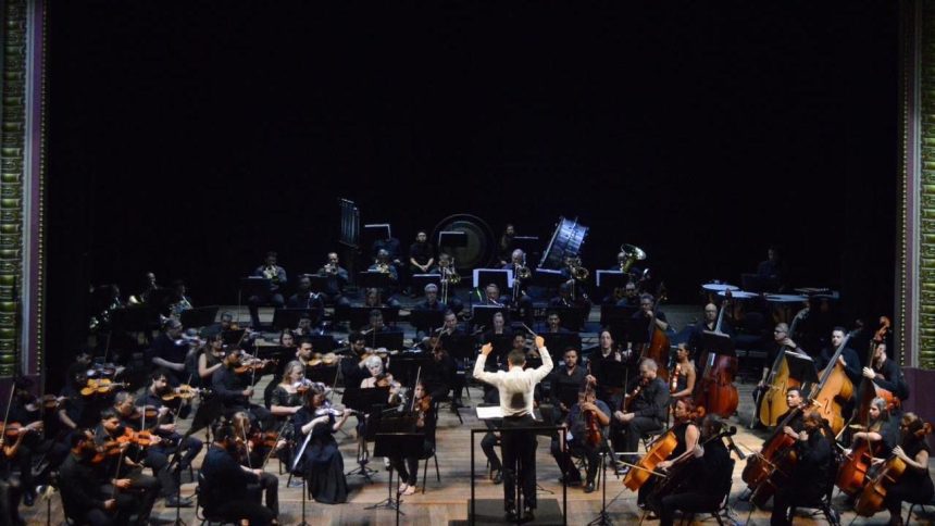 Orquestra sinfônica no palco, com maestro conduzindo e músicos tocando instrumentos.