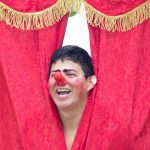 Palhaço sorrindo atrás de cortina vermelha em festival de teatro.