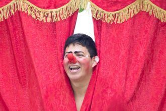 Palhaço sorrindo atrás de cortina vermelha em festival de teatro.