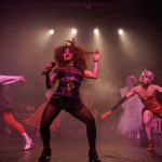 Performance drag no palco com luzes e figurinos elaborados, mostrando a palavra 'PATRICIA'