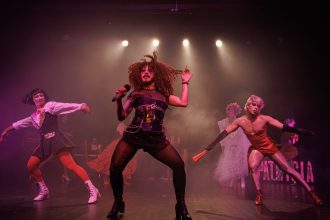 Performance drag no palco com luzes e figurinos elaborados, mostrando a palavra 'PATRICIA'