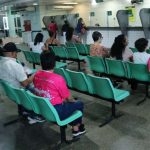 Pessoas sentadas em cadeiras em uma sala de espera, possivelmente em uma unidade de saúde.