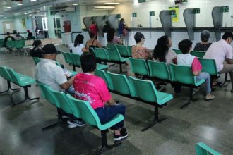 Pessoas sentadas em cadeiras em uma sala de espera, possivelmente em uma unidade de saúde.
