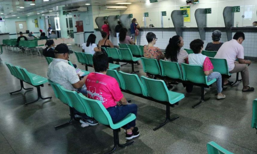 Pessoas sentadas em cadeiras em uma sala de espera, possivelmente em uma unidade de saúde.