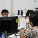 Pessoas trabalhando em escritório com computadores, possivelmente em processo seletivo ou prestando atendimento.