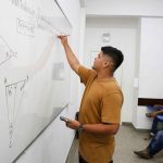 Professor escrevendo em quadro branco com alunos em sala de aula. Conteúdo de matemática.