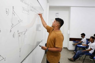 Professor escrevendo em quadro branco com alunos em sala de aula. Conteúdo de matemática.