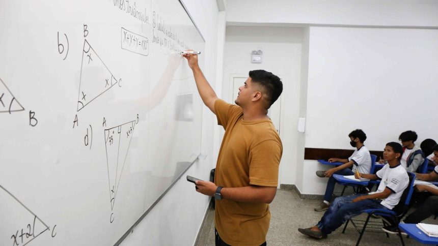 Professor escrevendo em quadro branco com alunos em sala de aula. Conteúdo de matemática.