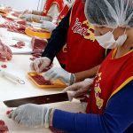 Profissionais preparando carne em açougue, com aventais e toucas.