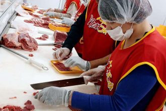 Profissionais preparando carne em açougue, com aventais e toucas.