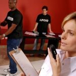Profissional fala ao celular e segura prancheta em evento do Senac, com equipe ao fundo.
