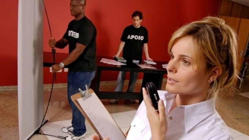 Profissional fala ao celular e segura prancheta em evento do Senac, com equipe ao fundo.