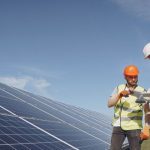 Técnicos analisando painéis solares em instalação, sob céu azul.