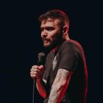 Tiago Santineli segurando um microfone em um palco escuro