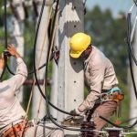 Trabalhadores da rede elétrica usando equipamentos de proteção individual (EPIs) em poste de energia.