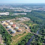 Vista aérea de tanques de armazenamento de combustíveis em meio à vegetação exuberante, com cidade ao fundo.