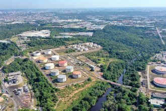Vista aérea de tanques de armazenamento de combustíveis em meio à vegetação exuberante, com cidade ao fundo.