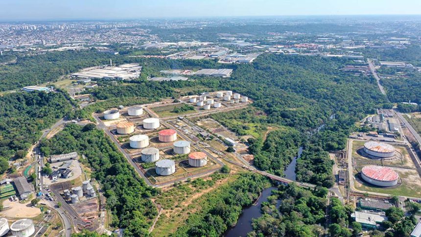 Vista aérea de tanques de armazenamento de combustíveis em meio à vegetação exuberante, com cidade ao fundo.