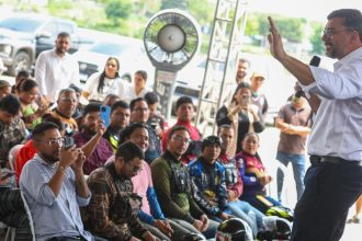Governador Wilson Lima discursa para multidão em evento ao ar livre, com pessoas registrando com celulares.