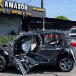 Carro destruído após acidente, com viatura policial ao fundo.