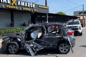 Carro destruído após acidente, com viatura policial ao fundo.