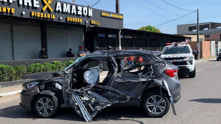 Carro destruído após acidente, com viatura policial ao fundo.