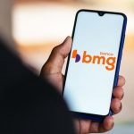 Celular mostrando logo do Banco BMG na mão de pessoa, com notícia sobre devolução de valores do INSS.