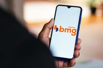 Celular mostrando logo do Banco BMG na mão de pessoa, com notícia sobre devolução de valores do INSS.