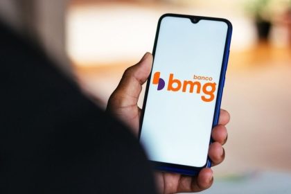 Celular mostrando logo do Banco BMG na mão de pessoa, com notícia sobre devolução de valores do INSS.