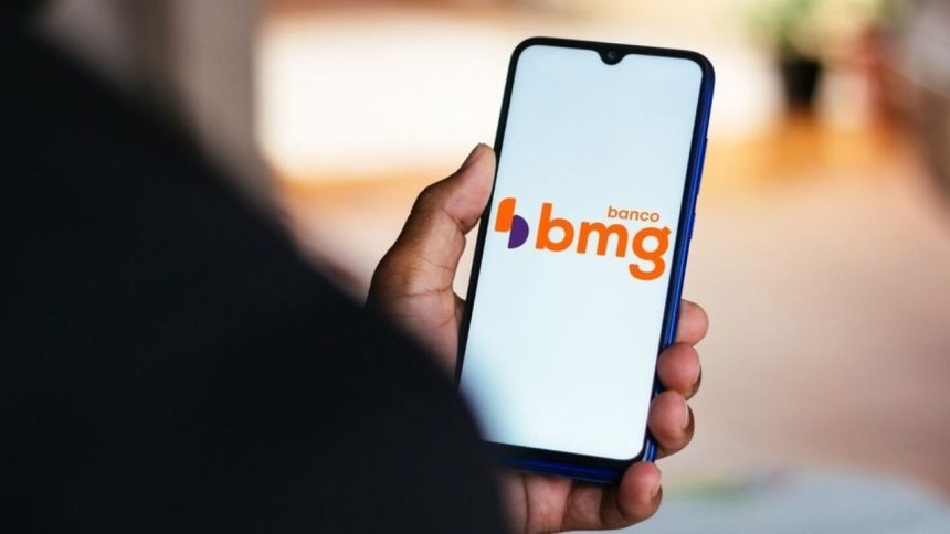 Celular mostrando logo do Banco BMG na mão de pessoa, com notícia sobre devolução de valores do INSS.