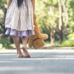 Criança caminha descalça com urso de pelúcia em um parque, aludindo à infância e vulnerabilidade.