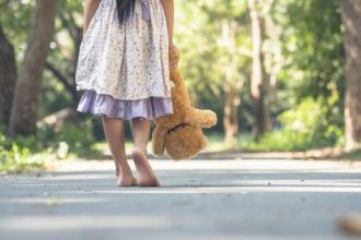 Criança caminha descalça com urso de pelúcia em um parque, aludindo à infância e vulnerabilidade.