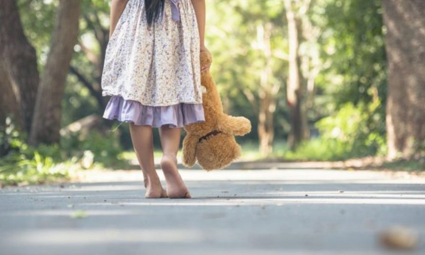 Criança caminha descalça com urso de pelúcia em um parque, aludindo à infância e vulnerabilidade.