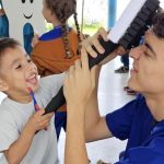 Criança sorrindo escovando os dentes com a ajuda de um profissional em projeto de saúde bucal.