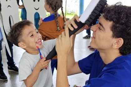 Criança sorrindo escovando os dentes com a ajuda de um profissional em projeto de saúde bucal.